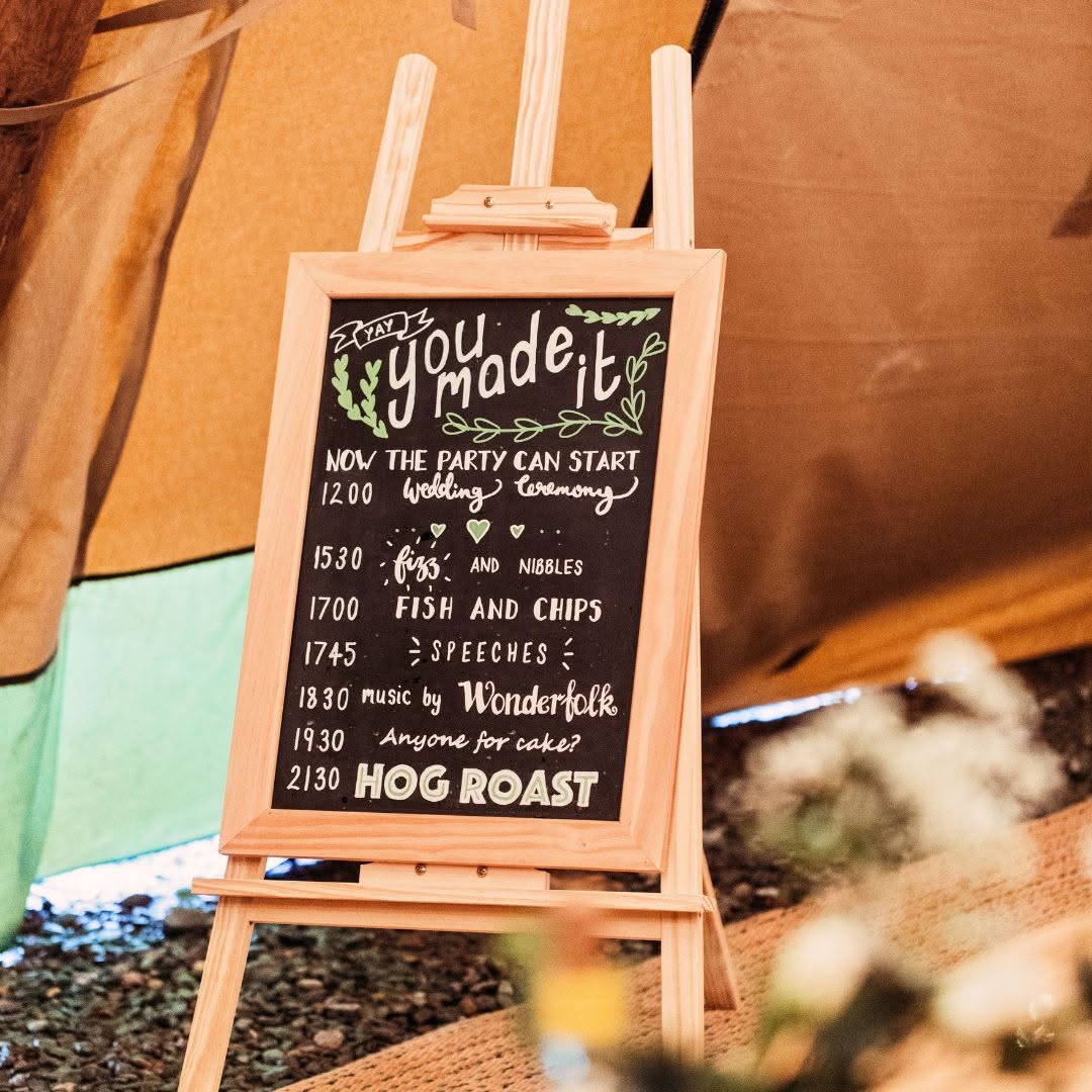 Handwritten chalkboard on an easel at a tipi entrance, greeting guests and listing the running order